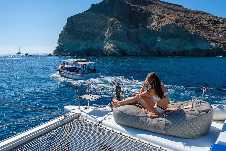 The catamaran's bow off the Akrotiri headland at Mesa Pigadia, deep blue water below the cliffs